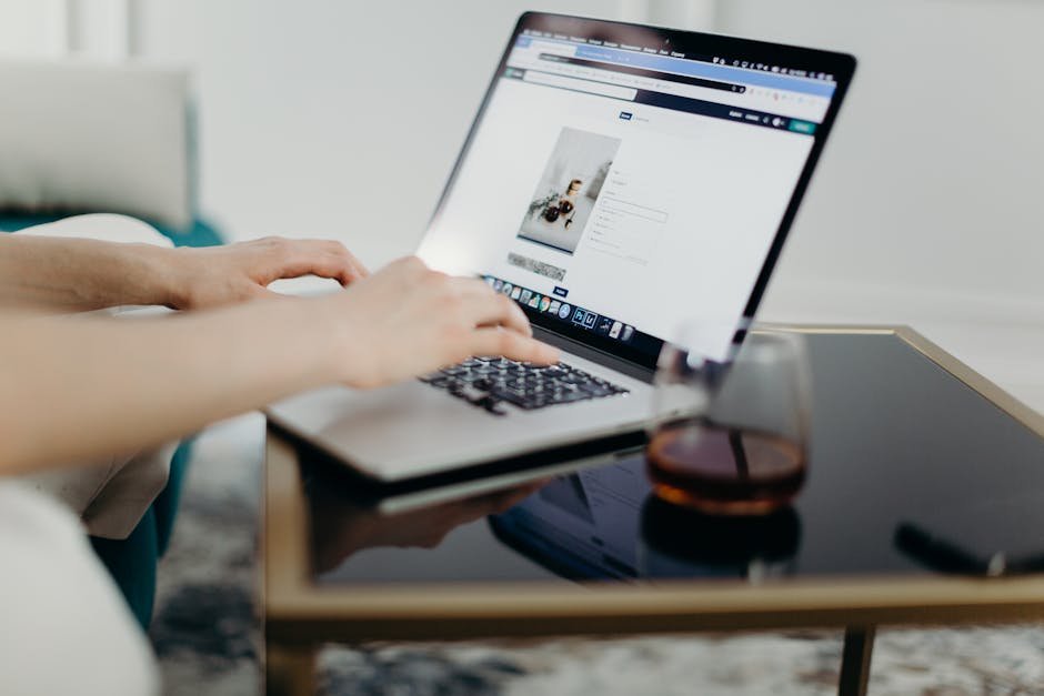 A person typing on a laptop in a stylish home office with a glass of drink nearby.